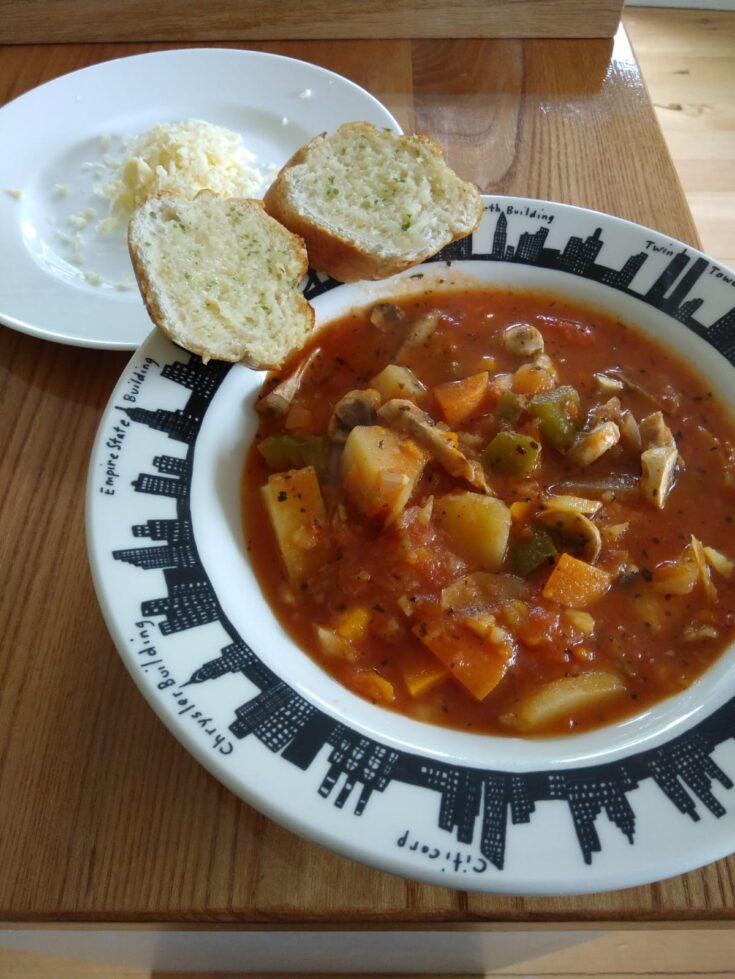 A bowl of simple soup served with two slices of garlic bread. Chunks of potato, carrot and green pepper can be seen against a tomatoey liquid base.